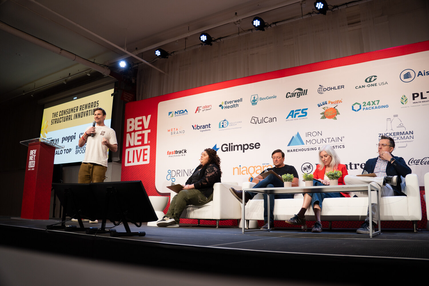 Presenter in a cream T-shirt stands under a slide naming Oatly, poppi, Chobani and Halo Top while four judges sit on couches.