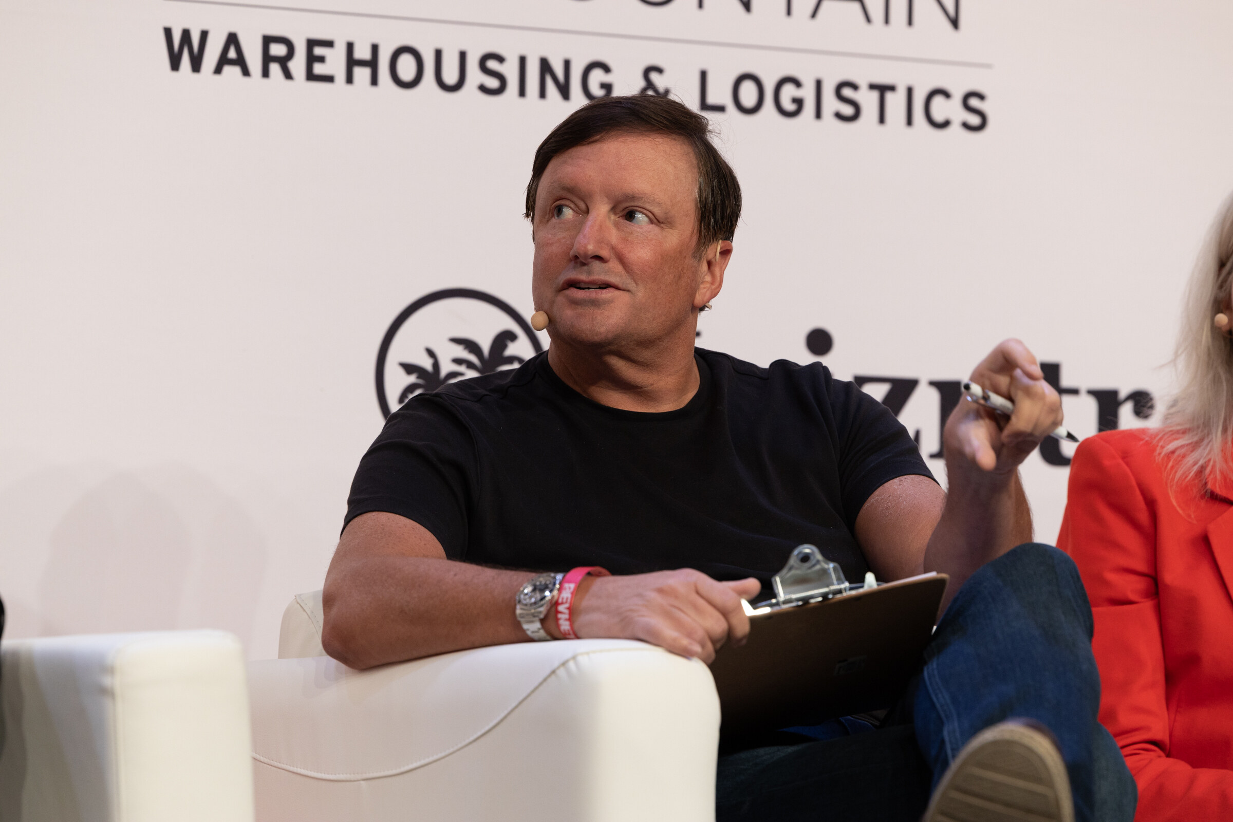 Man in a black T-shirt sits on a white couch with a clipboard, looking toward the stage.