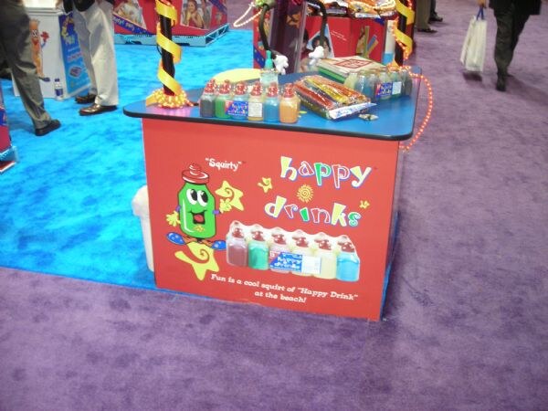 A red Happy Drinks display counter with a green bottle cartoon character and a row of squeezable drink bottles, standing on blue and purple carpet.