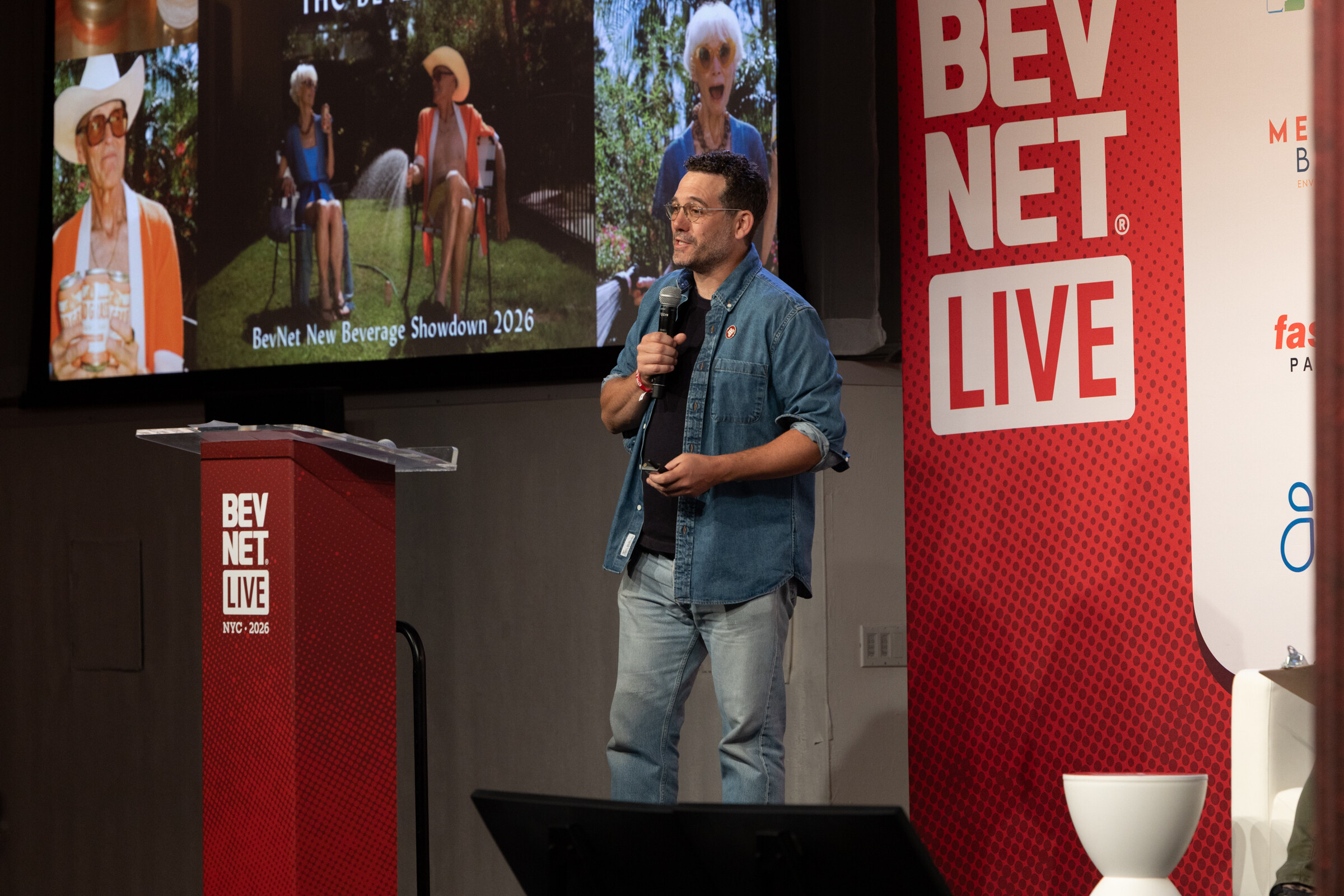 Man in a denim shirt speaks into a microphone in front of a Dad Grass garden-party slide labeled BevNet New Beverage Showdown 2026.