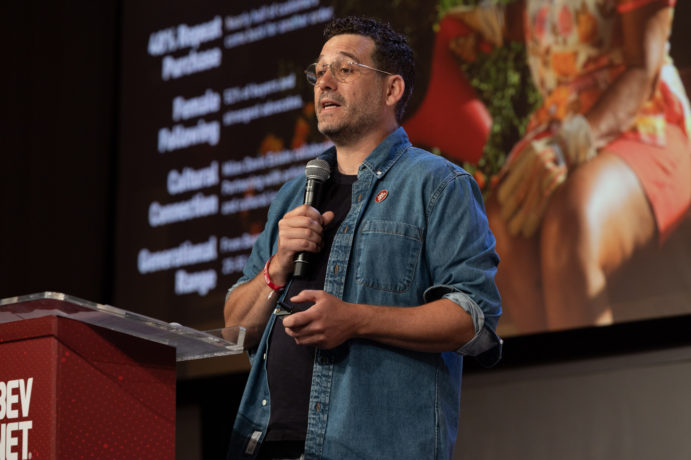 Man in glasses and a denim shirt speaks into a microphone in front of a slide about repeat purchases.