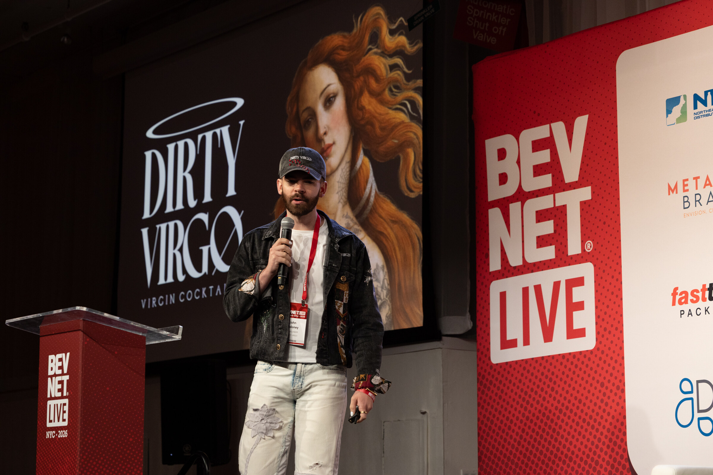 Man in a cap and denim jacket speaks into a microphone in front of a Dirty Virgo Virgin Cocktails slide.