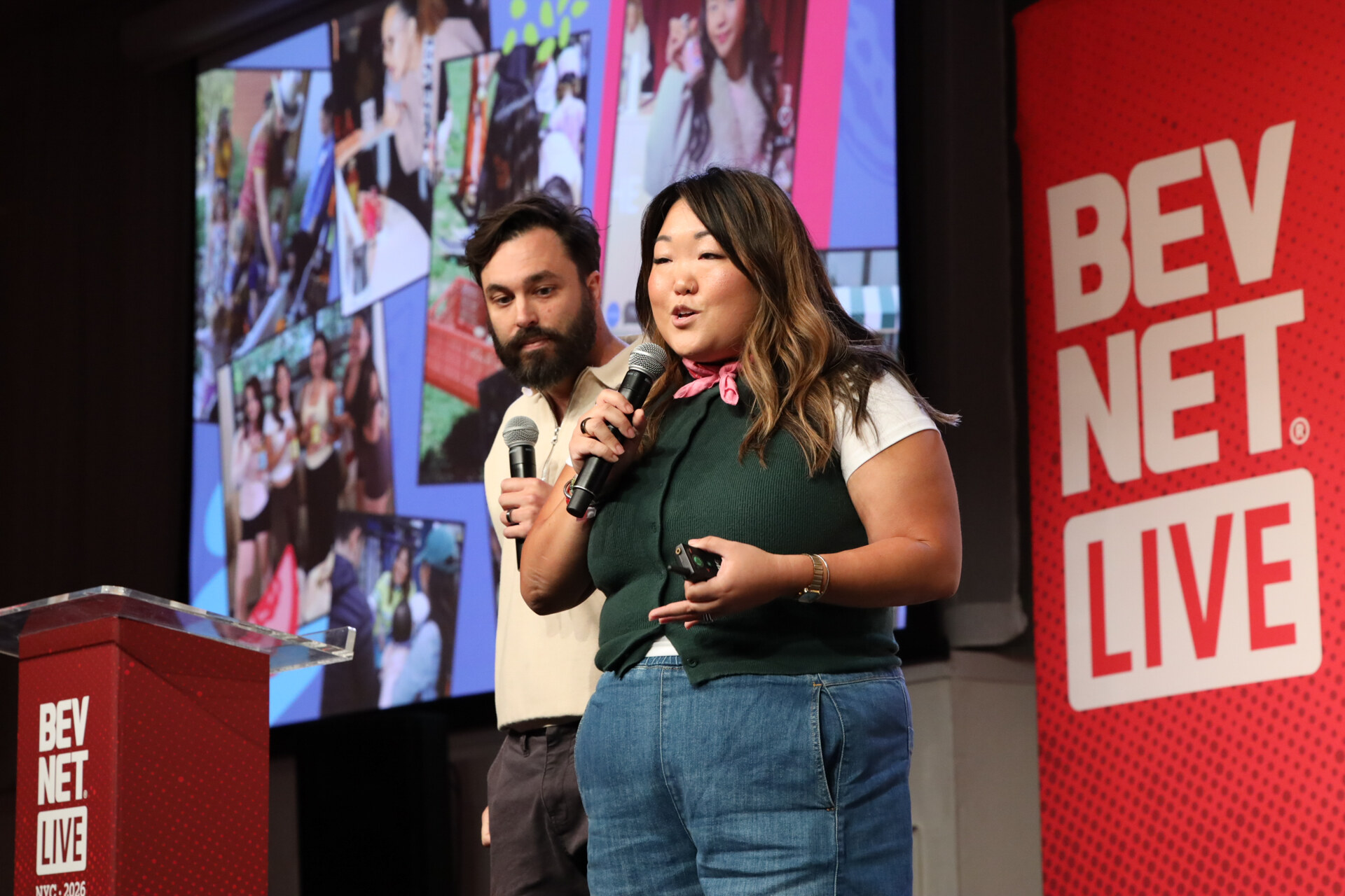 Woman in a green top and pink neckerchief speaks into a microphone beside a man, in front of a collage slide.