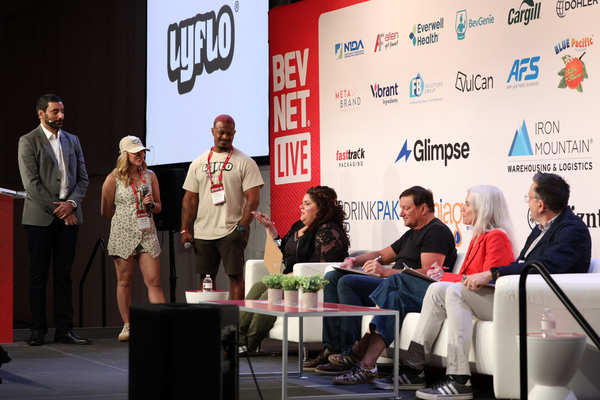 Host and two Lyflo presenters stand on stage beside four judges on couches, with a Lyflo logo on screen.
