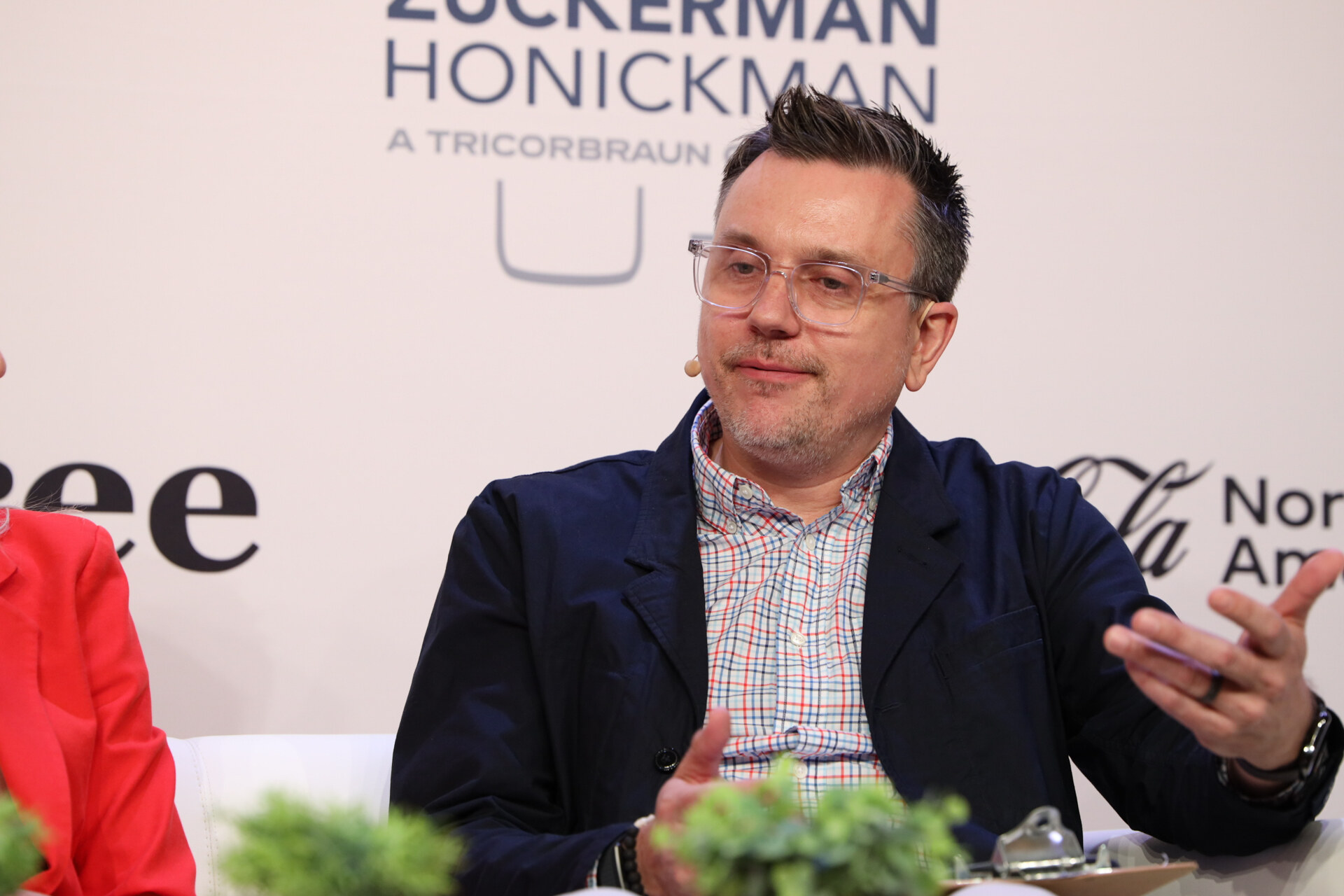 Man in clear glasses, a plaid shirt and navy jacket gestures while speaking from a couch.