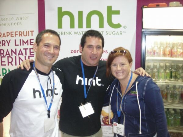 Two men and a woman pose with arms around each other in front of a Hint banner. The men wear Hint shirts, and all three wear blue lanyards with badges. A drink cooler is at right.