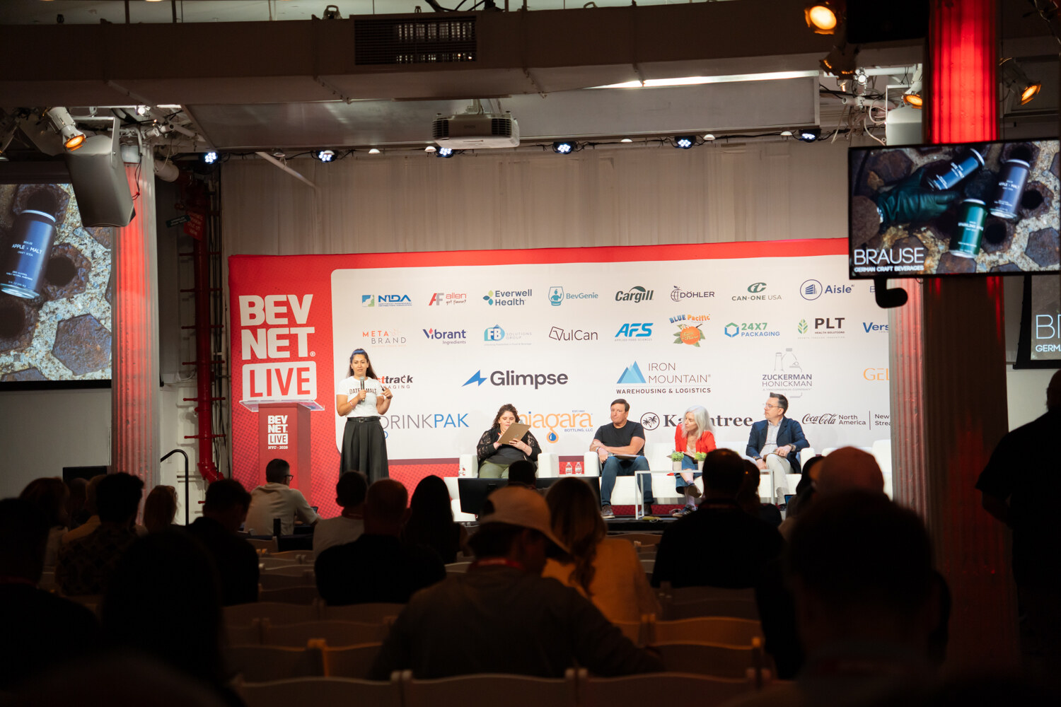 Wide view over a seated audience toward a red BevNET Live stage, with a presenter, four judges and a Brause slide on screen.