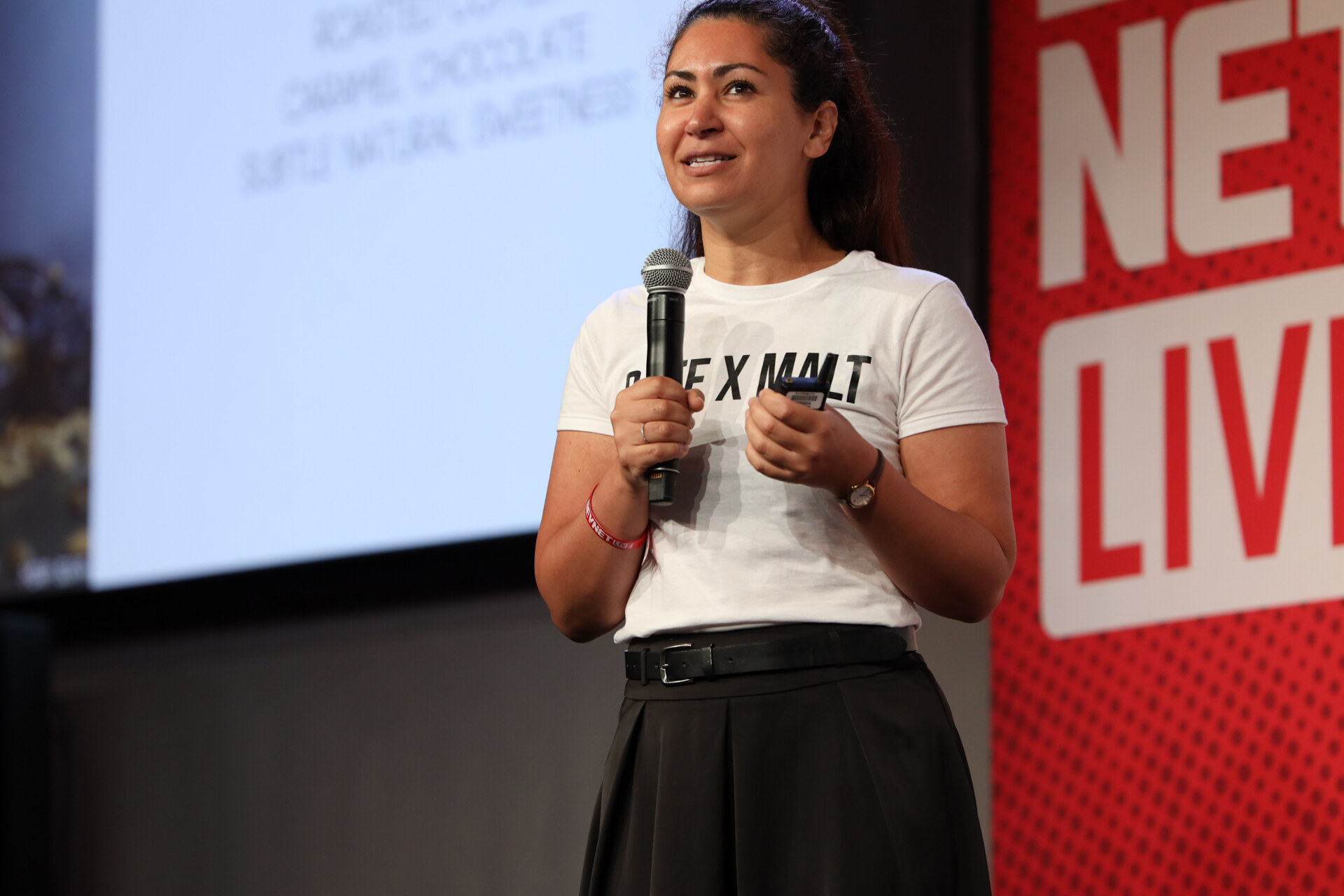 Smiling woman in a Date x Malt T-shirt and black skirt speaks into a microphone on stage.