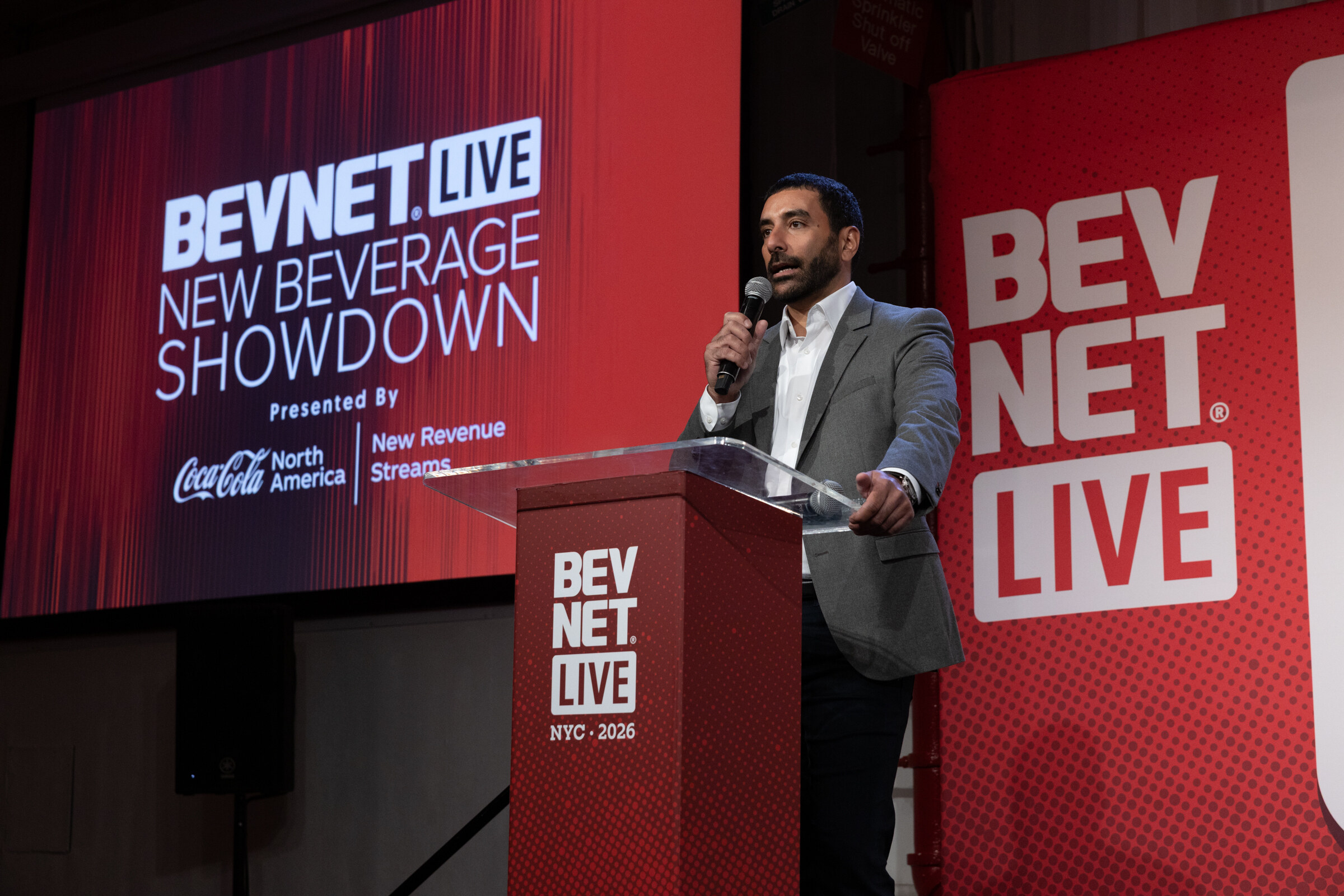 Man in a grey jacket speaks at a BevNET Live NYC 2026 podium in front of a red New Beverage Showdown screen.