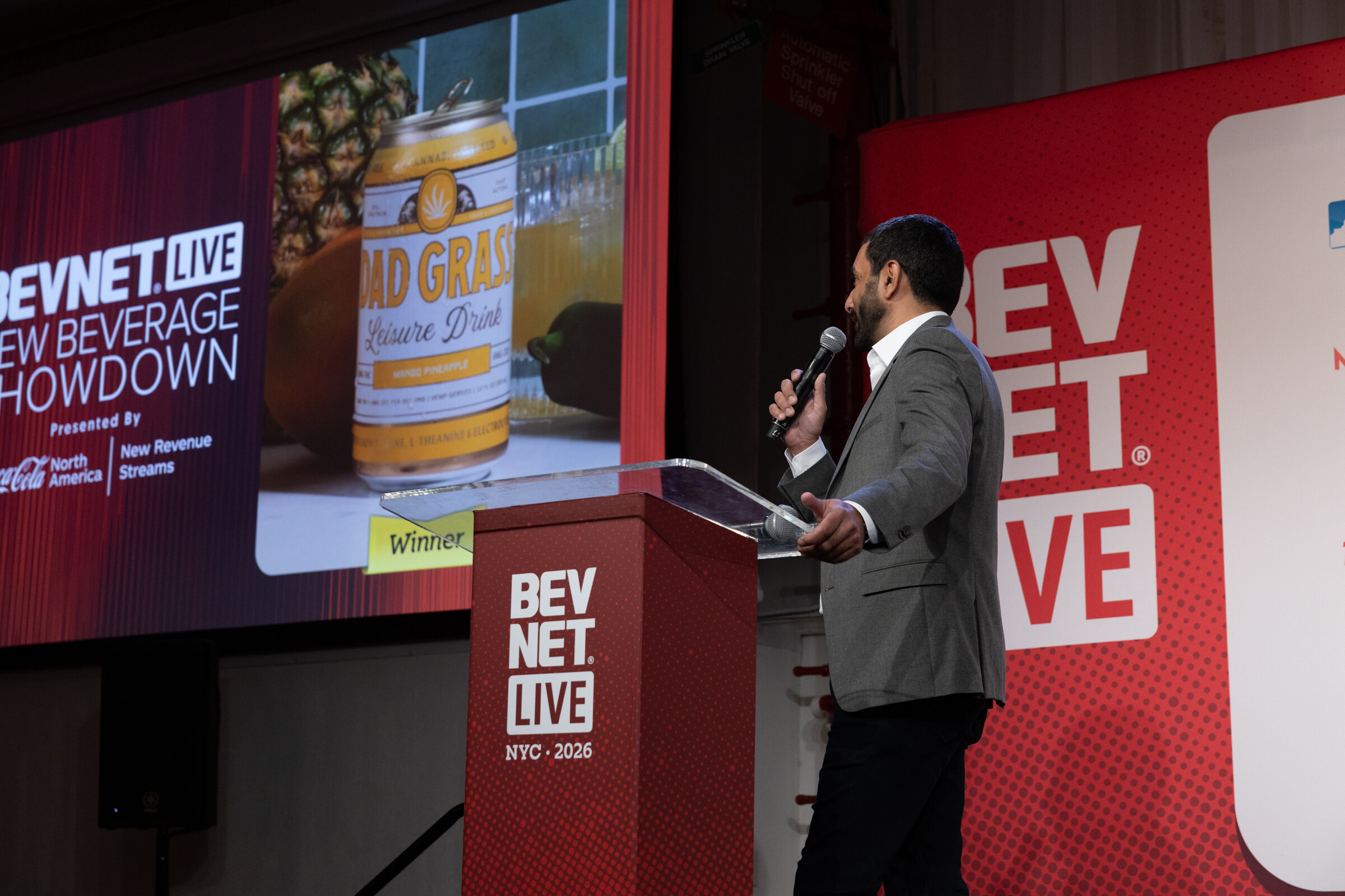 Screen shows a can of Dad Grass Leisure Drink with a Winner label beside the New Beverage Showdown logo as the host speaks at the podium.