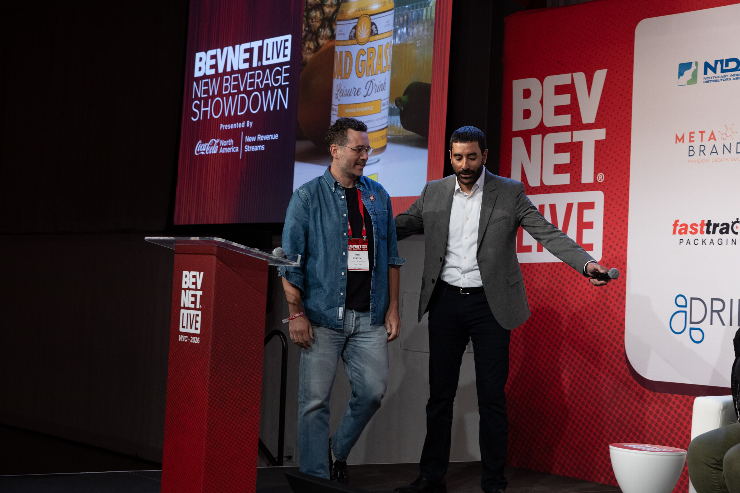 Man in a denim shirt walks on stage beside the host in a grey jacket, with the New Beverage Showdown screen behind them.