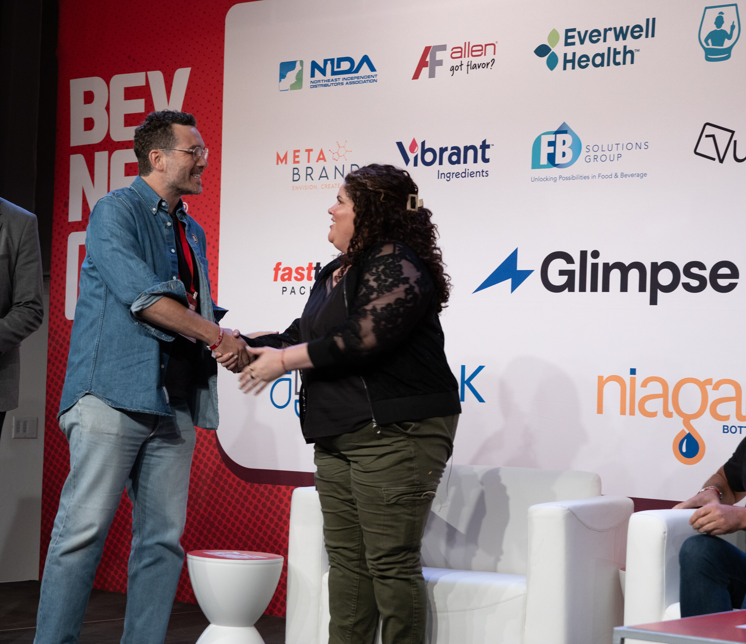 Man in a denim shirt shakes hands with a woman in black on stage in front of the sponsor wall.