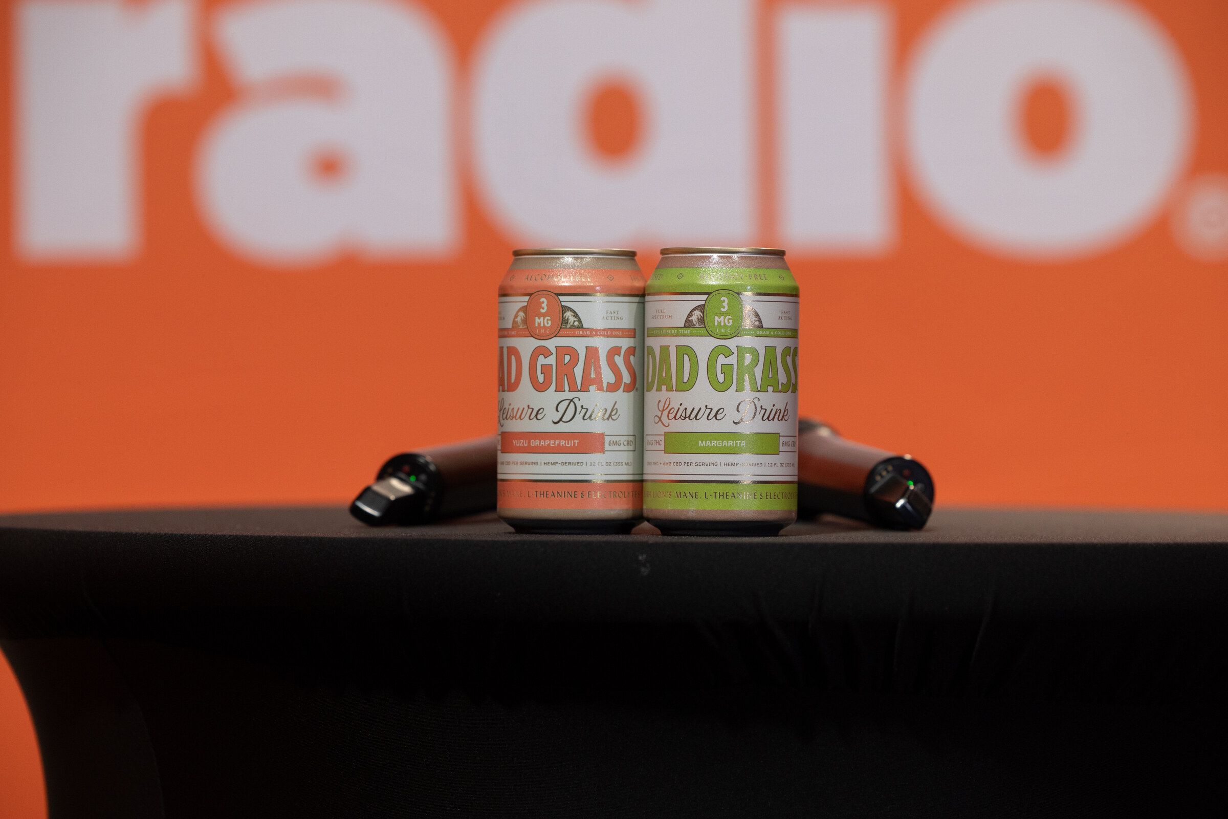 Two cans of Dad Grass Leisure Drink sit between microphones on a black table in front of an orange Taste Radio backdrop.