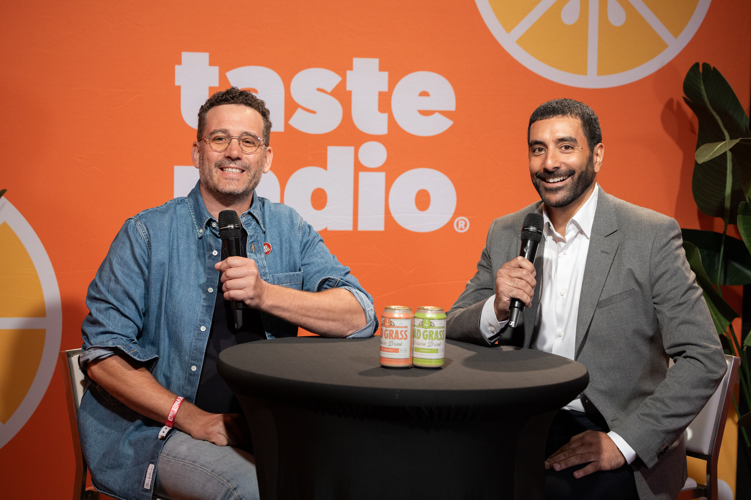 Two smiling men with microphones sit at a table with Dad Grass cans in front of an orange Taste Radio backdrop.