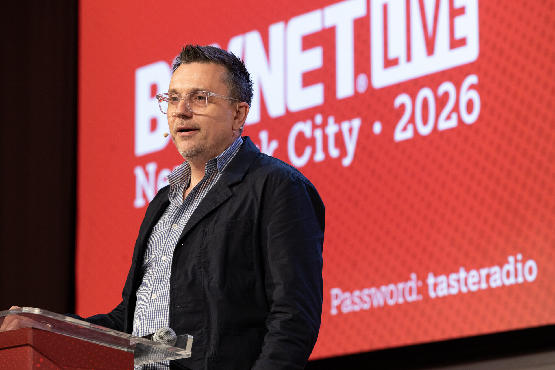 Man in glasses and a dark jacket speaking at a clear podium in front of a red screen reading BevNET Live New York City 2026.