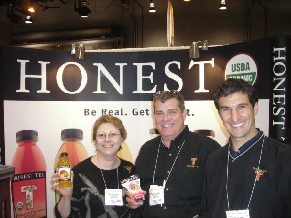 A woman and two men smile at an Honest Tea booth. The woman holds a bottle of tea and the man beside her holds a snack package. Honest Tea bottles and a USDA Organic logo are on the backdrop.