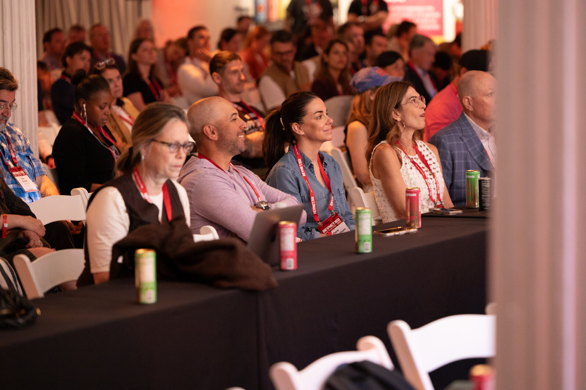 Attendees wearing red lanyards seated at long tables with cans of drinks, watching the stage.