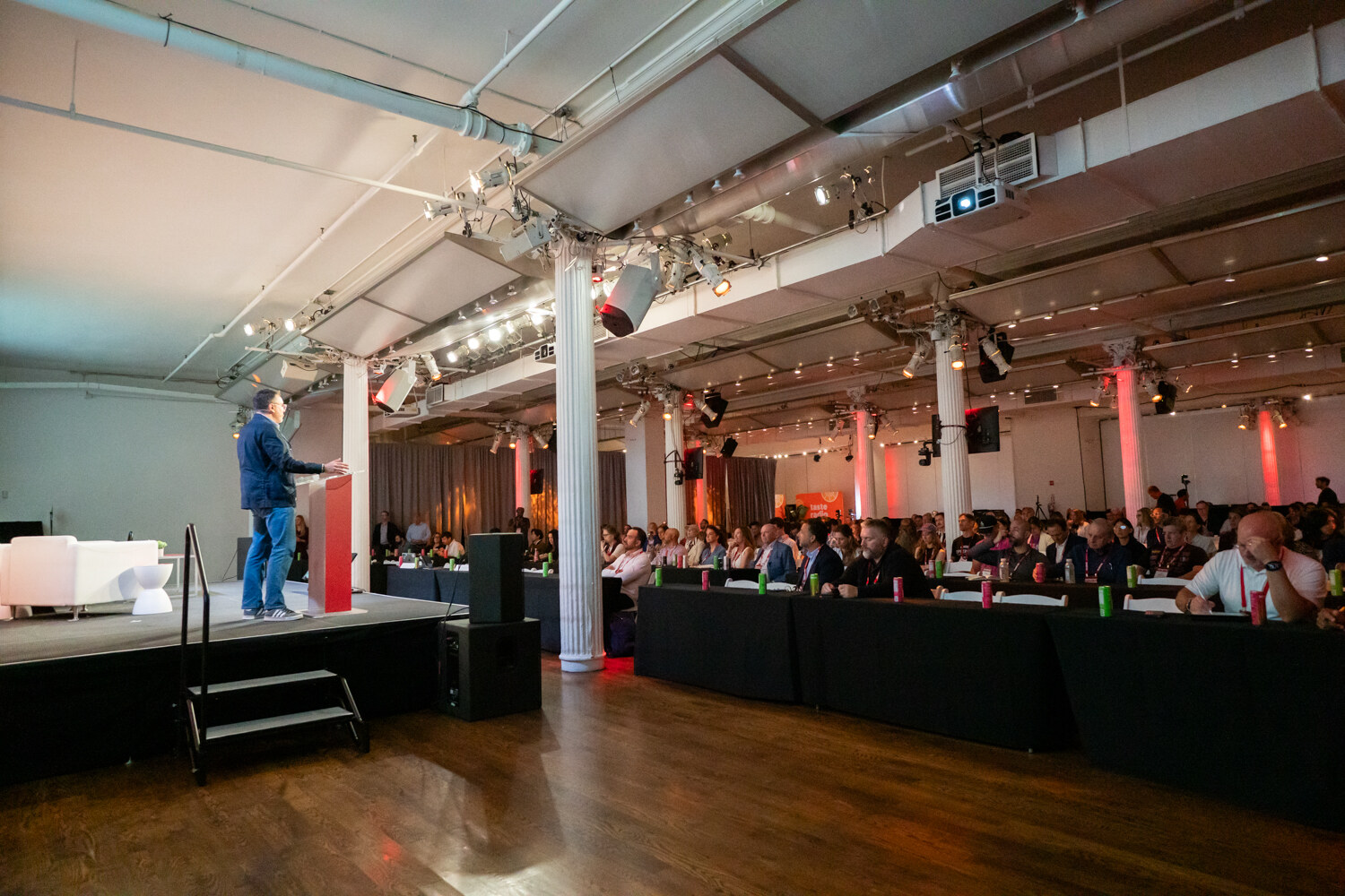 Wide view of a speaker at a podium on a stage at left, facing rows of attendees seated at long tables in a pillared hall.