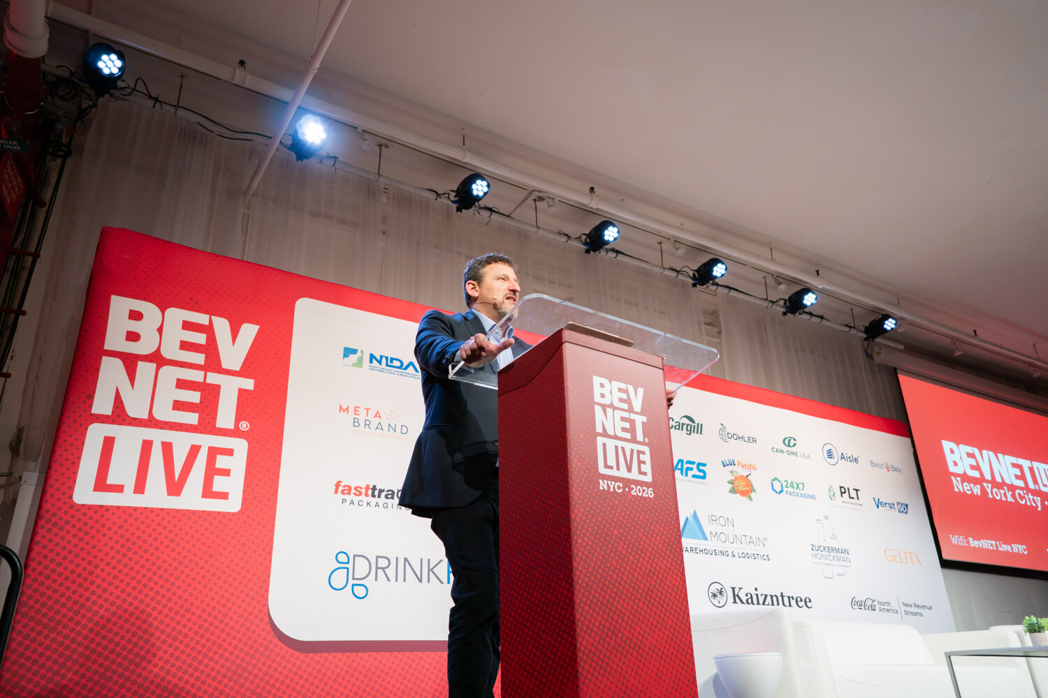 Low-angle view of a man in a navy blazer speaking at a red BevNET Live podium in front of a sponsor wall.