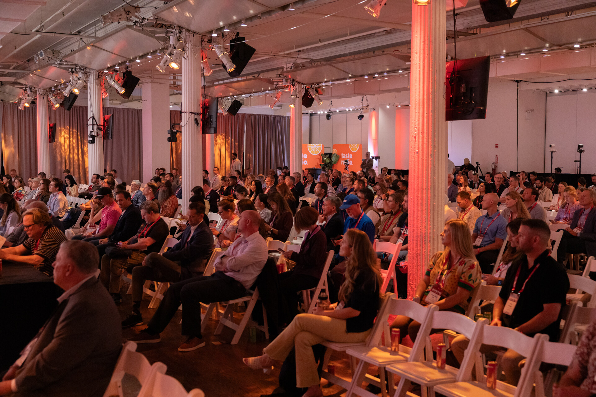 Rows of attendees on white folding chairs in a large pillared hall lit with warm stage lighting.