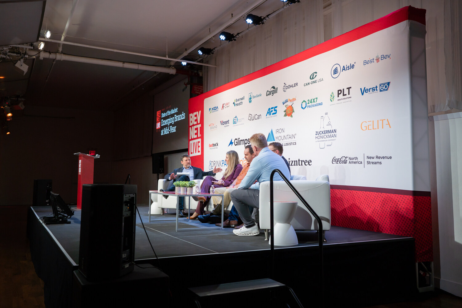 Side view of five people seated on white couches on a stage in front of a large sponsor wall.