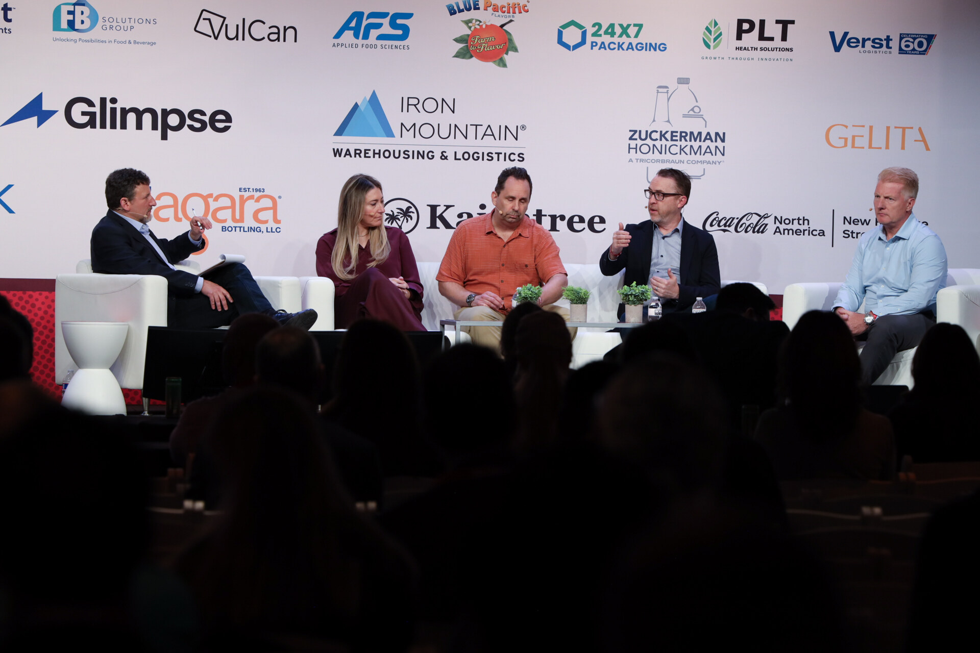 Five panelists seated across a stage in front of a sponsor wall, with audience silhouettes in the foreground.