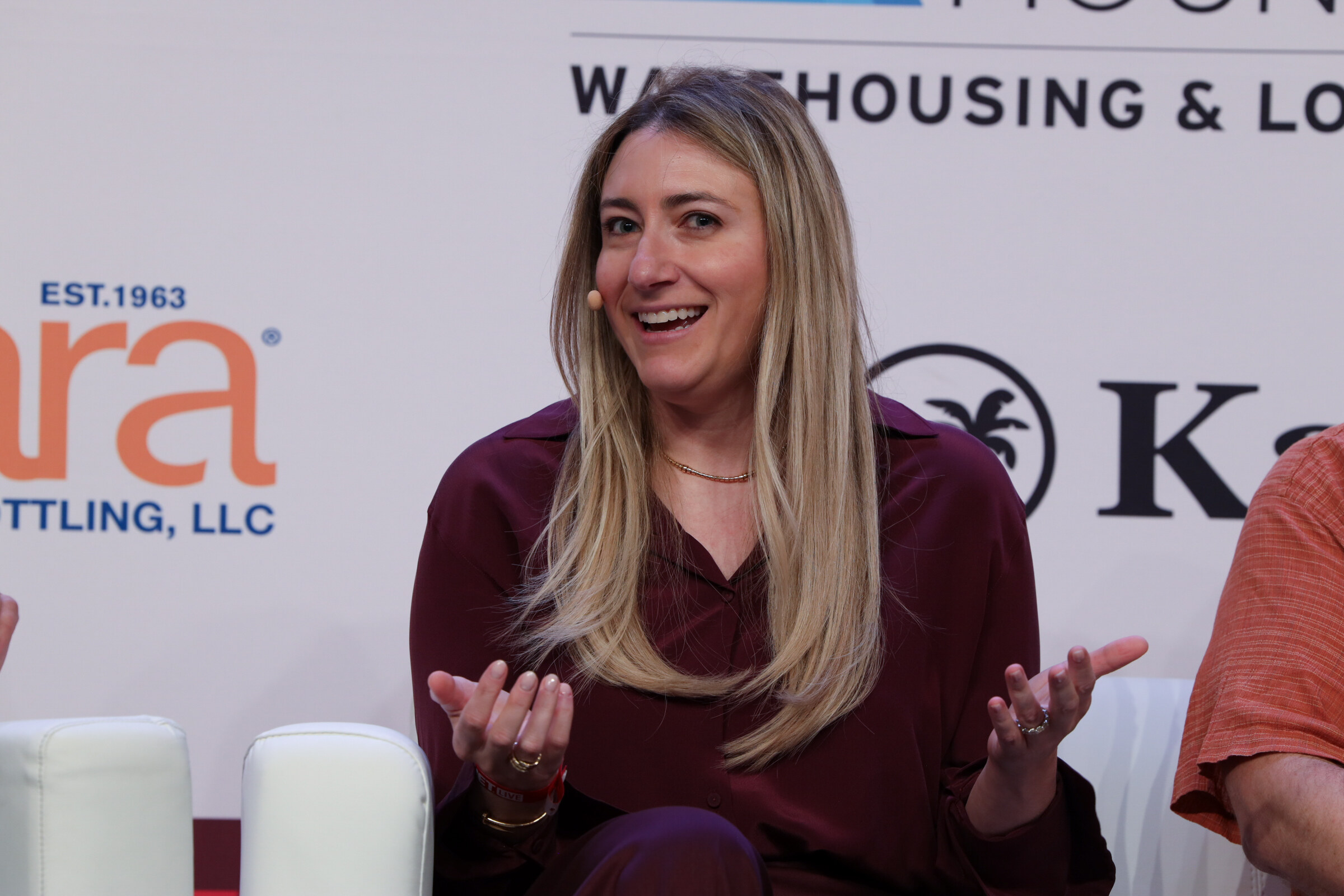 Smiling woman with long blond hair in a burgundy blouse gesturing with open hands while speaking on stage.