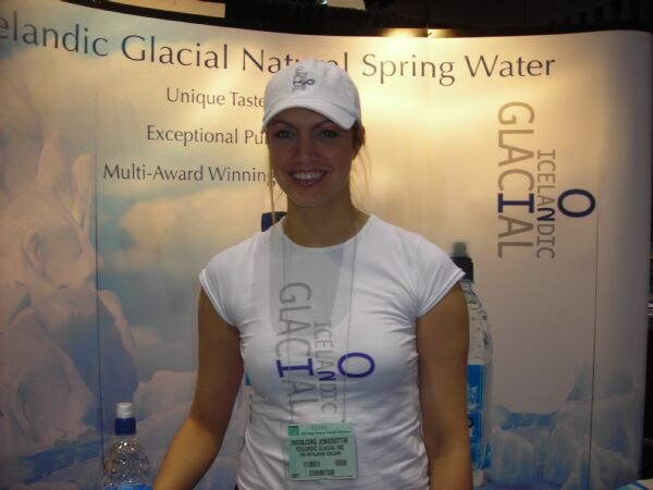 A smiling woman in a white cap and white Icelandic Glacial T-shirt stands in front of an Icelandic Glacial Natural Spring Water backdrop. She wears an exhibitor badge, and bottles are nearby.