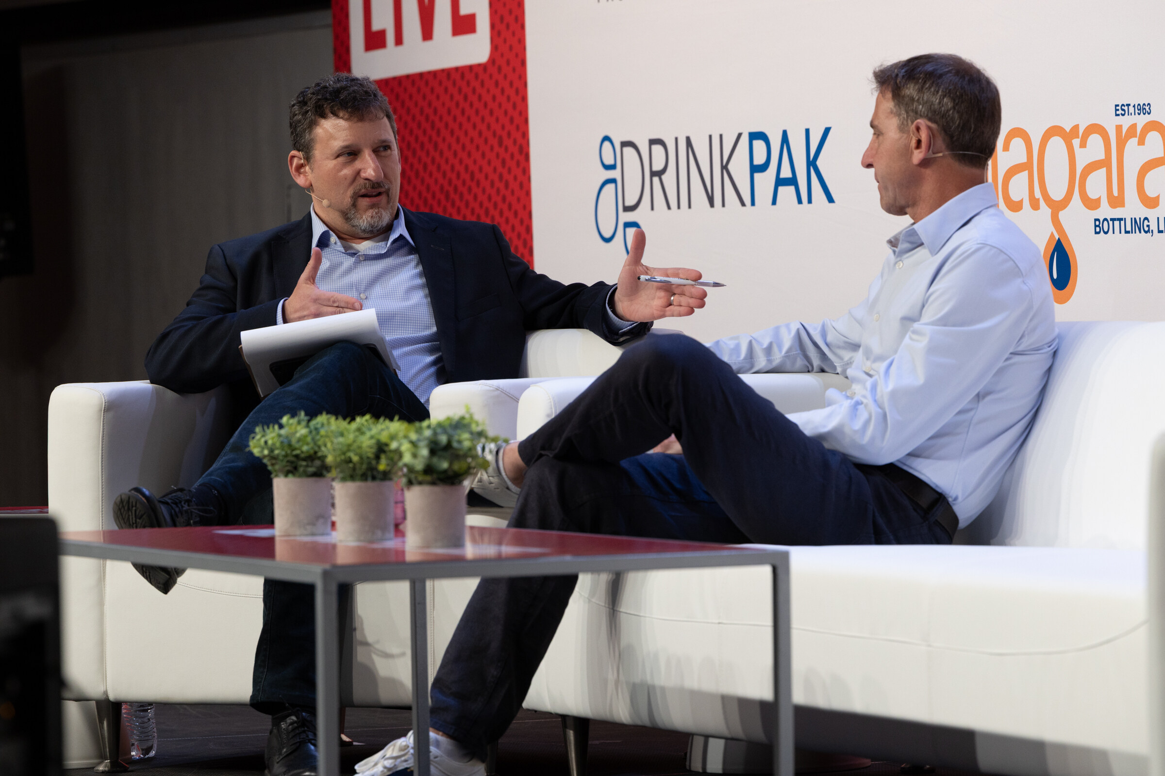 Two men in conversation on white couches on stage, one holding notes and gesturing.