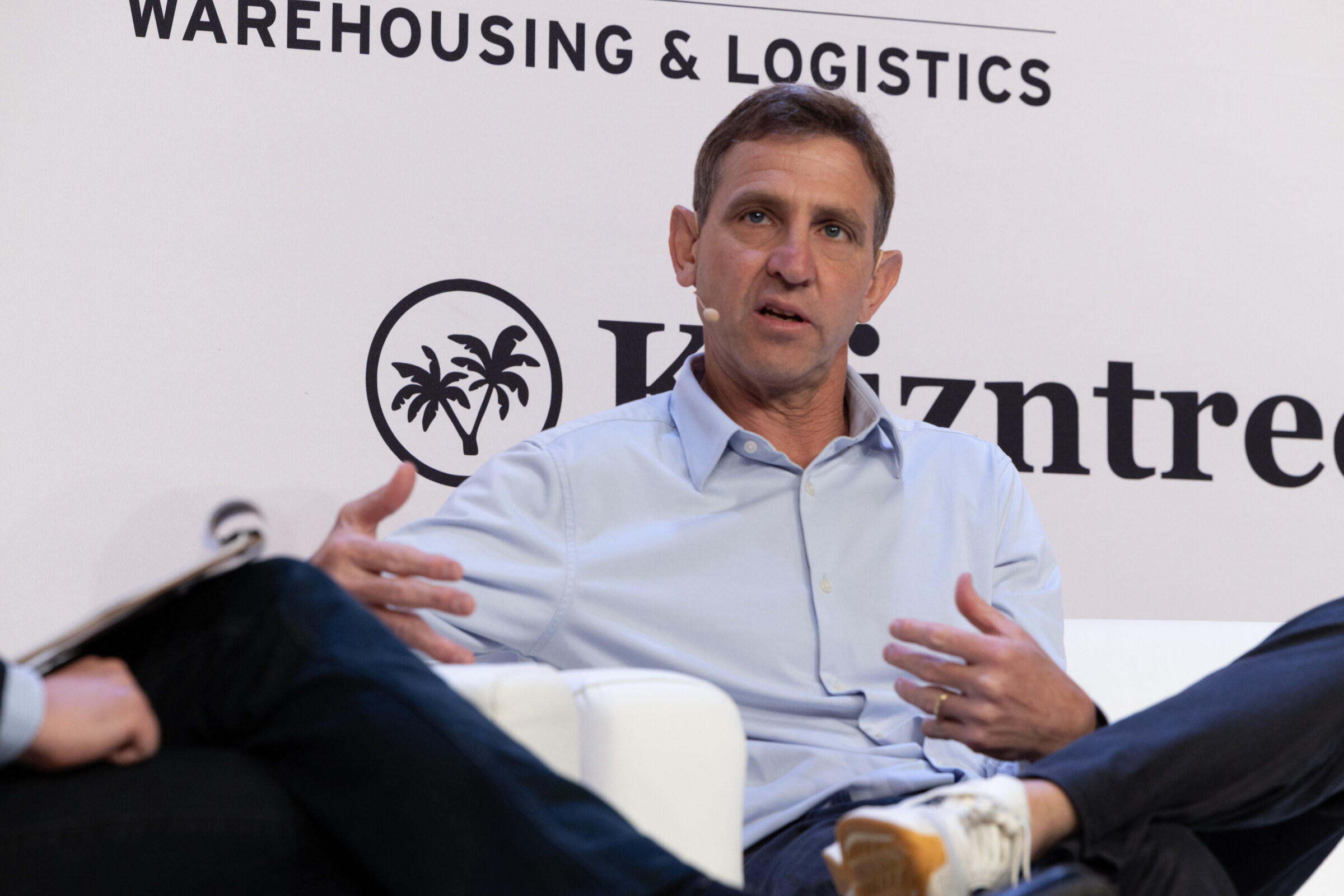 Man in a light blue shirt gesturing as he speaks from a white couch in front of a Kaizntree logo.