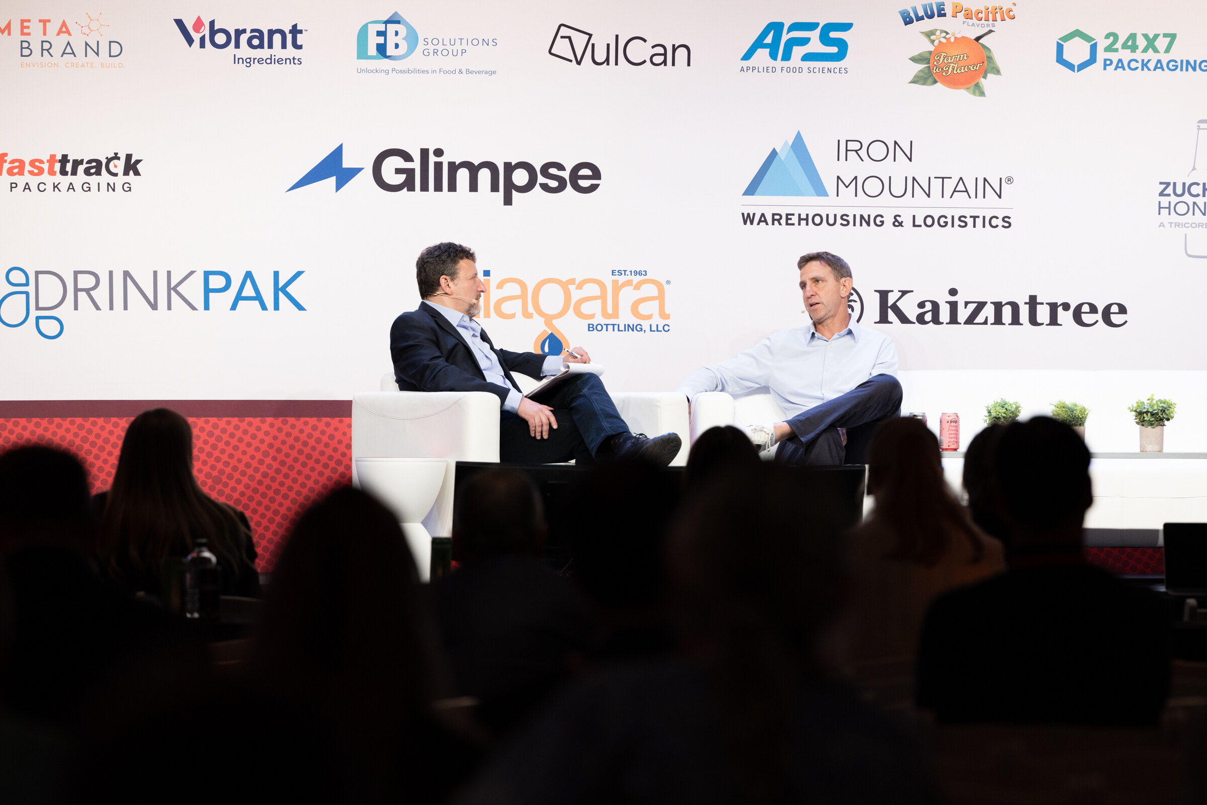 Two men seated on a stage in front of a sponsor wall, with audience silhouettes in the foreground.