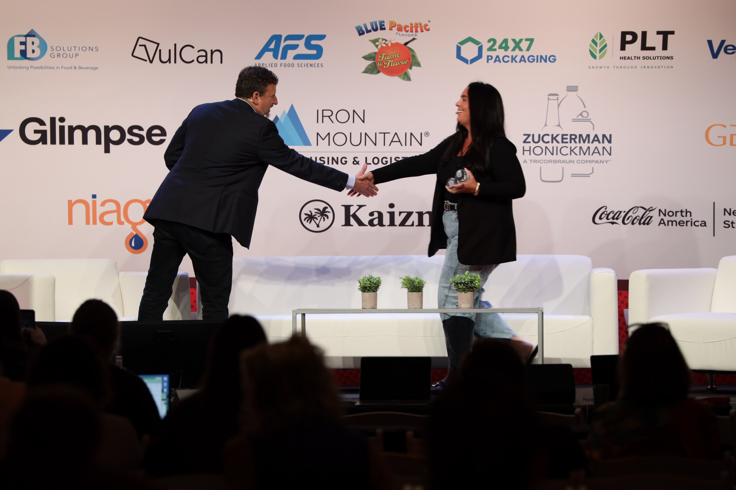 Man in a dark suit shaking hands with a woman in a black jacket and jeans on stage in front of a sponsor wall.