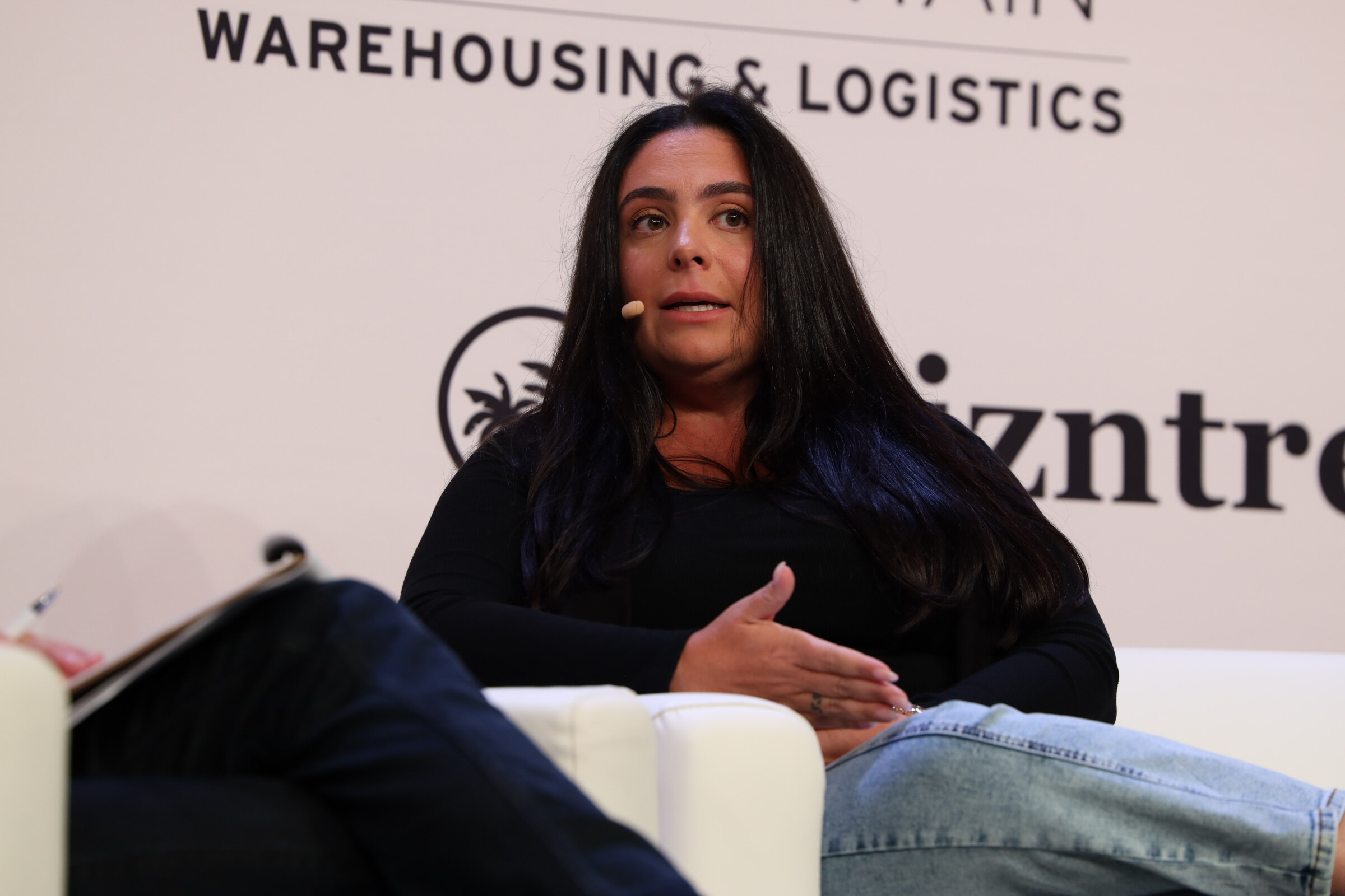 Woman with long dark hair speaking from a white couch on stage, one hand raised.