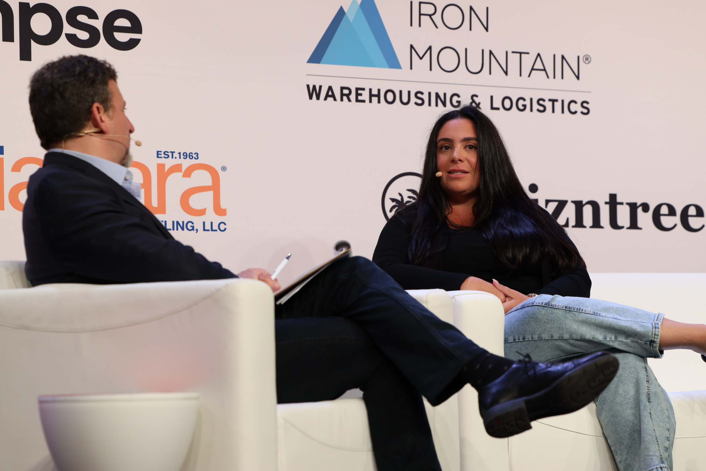 Man in a dark suit and a woman in a black top seated on white couches in conversation, in front of an Iron Mountain logo.
