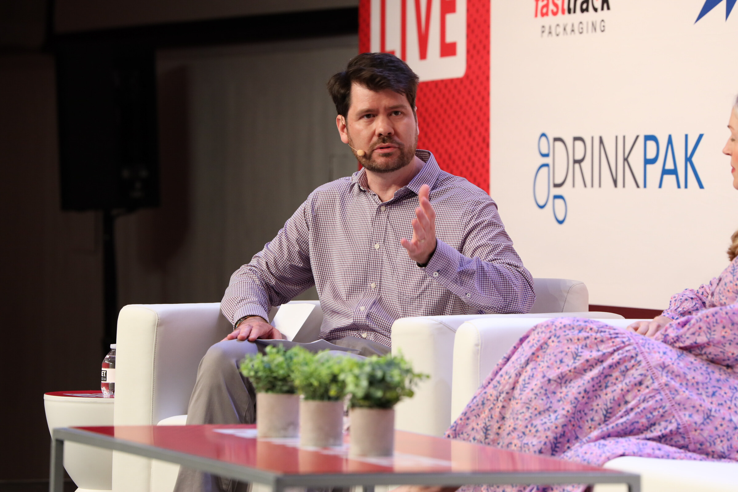 Man in a checked shirt gesturing while speaking from a white couch on stage.