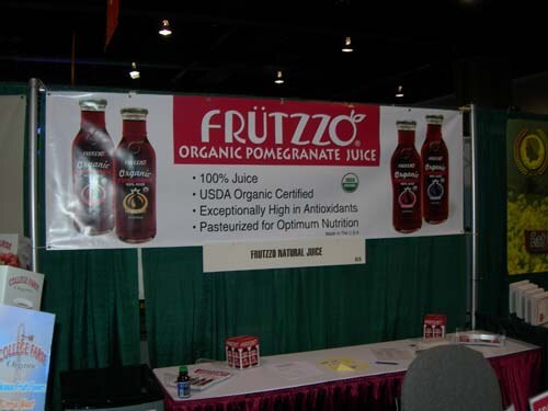 A booth with a banner reading Frützzo Organic Pomegranate Juice, flanked by bottle images, above a table with a red skirt holding small items and a bottle.
