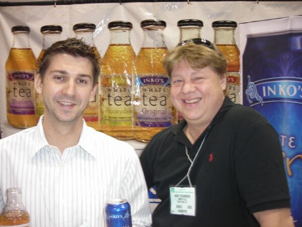 Two smiling men stand in front of a row of Inko's White Tea bottles in several flavors. The man at right wears a black polo, sunglasses on his head and an exhibitor badge. The man at left holds a can.