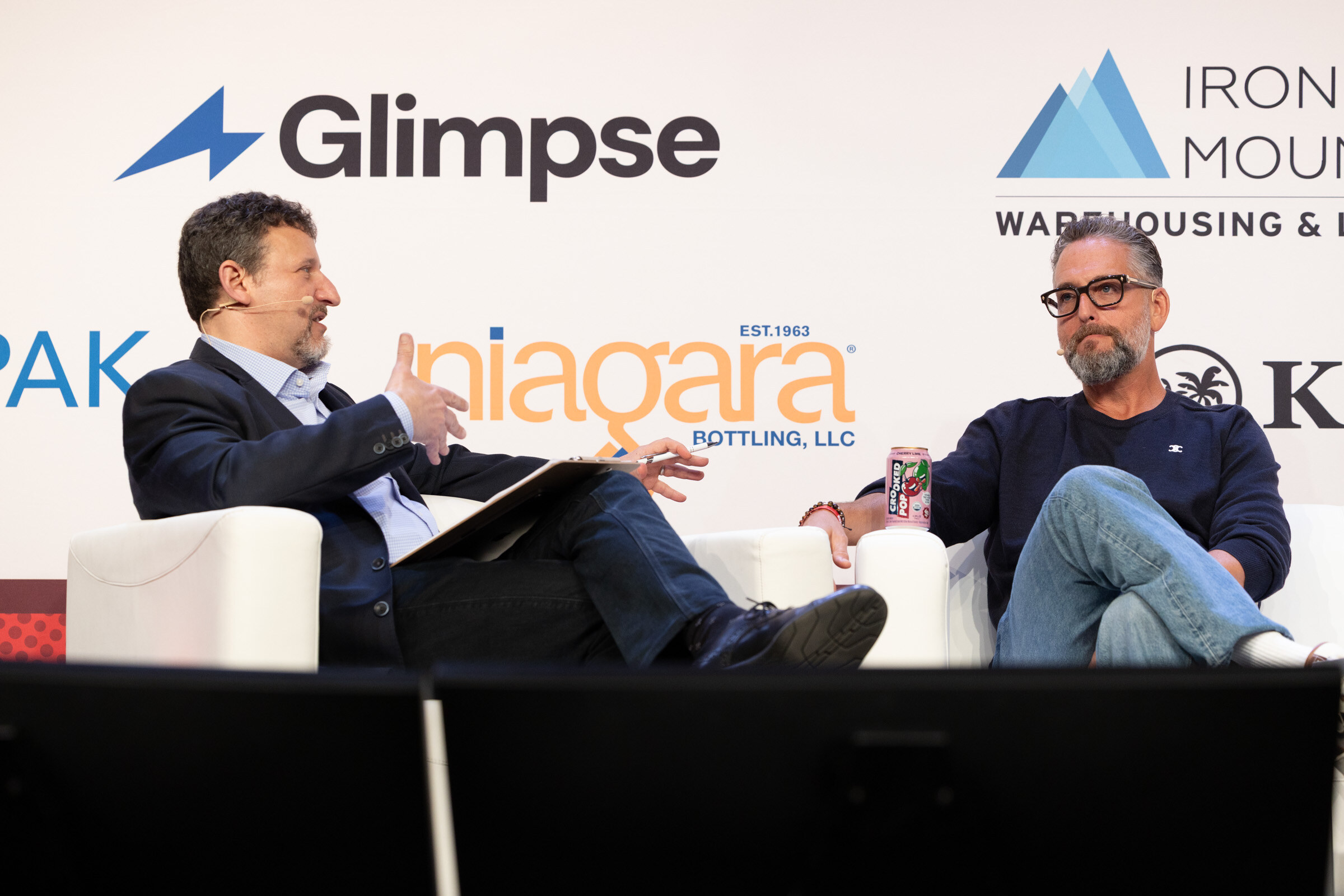 Man in a dark suit with notes talking with a man in glasses and a navy sweater on white couches in front of a Niagara Bottling logo.
