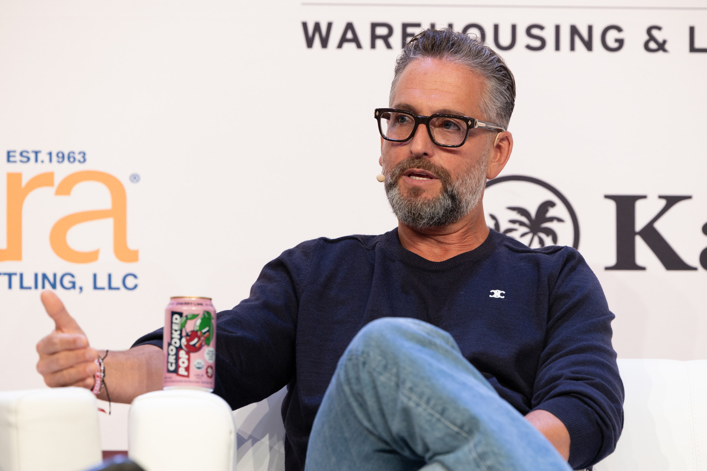 Man in black glasses and a navy sweater speaking from a couch, holding a pink can of Crooked Pop.