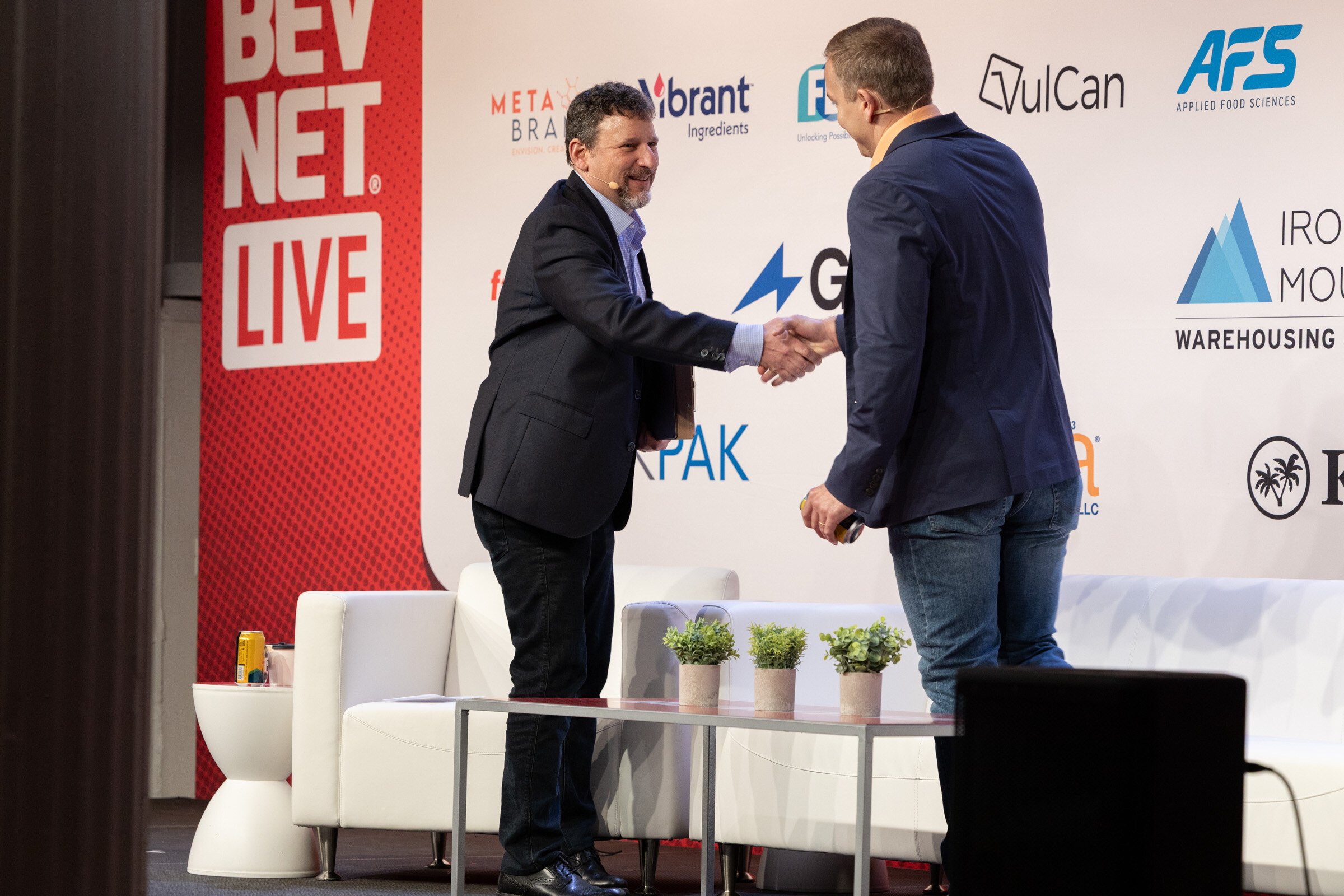 Two men shaking hands on stage beside white couches in front of a red BevNET Live banner.