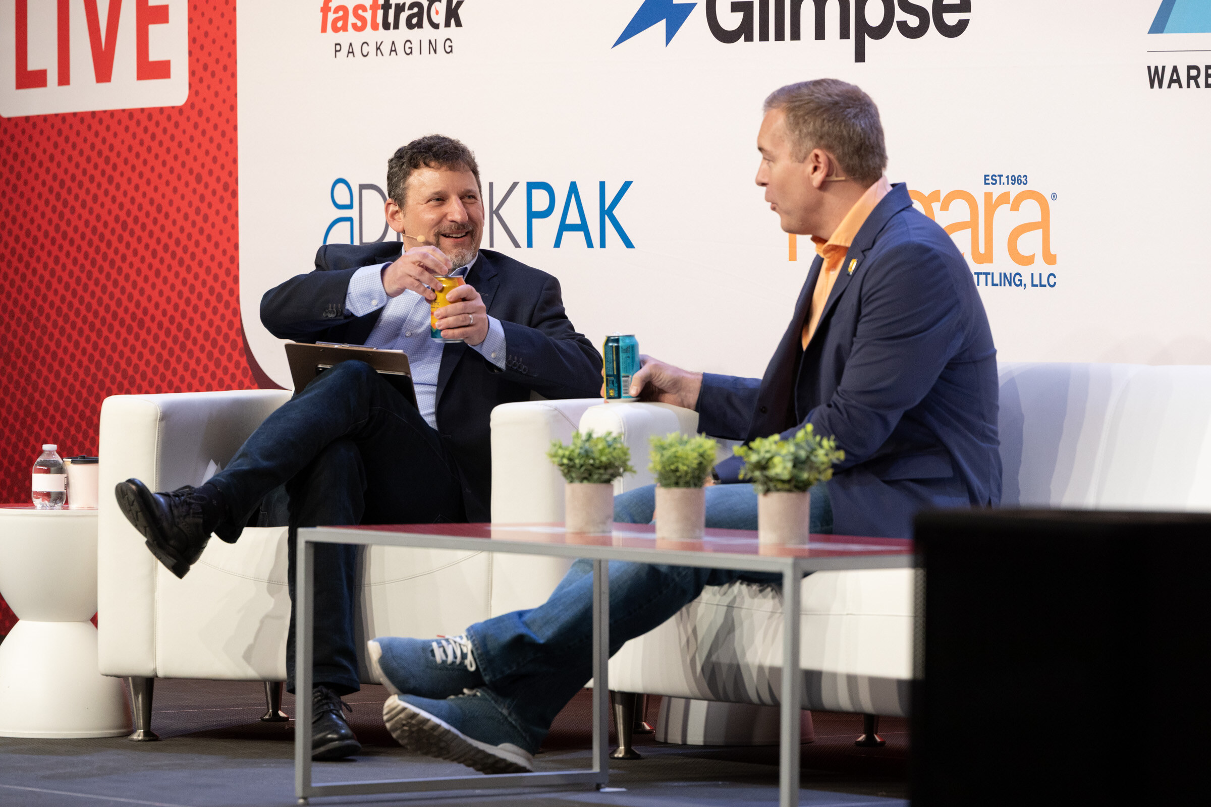 Two men on white couches laughing, one holding up a can, in front of DrinkPAK and Niagara logos.