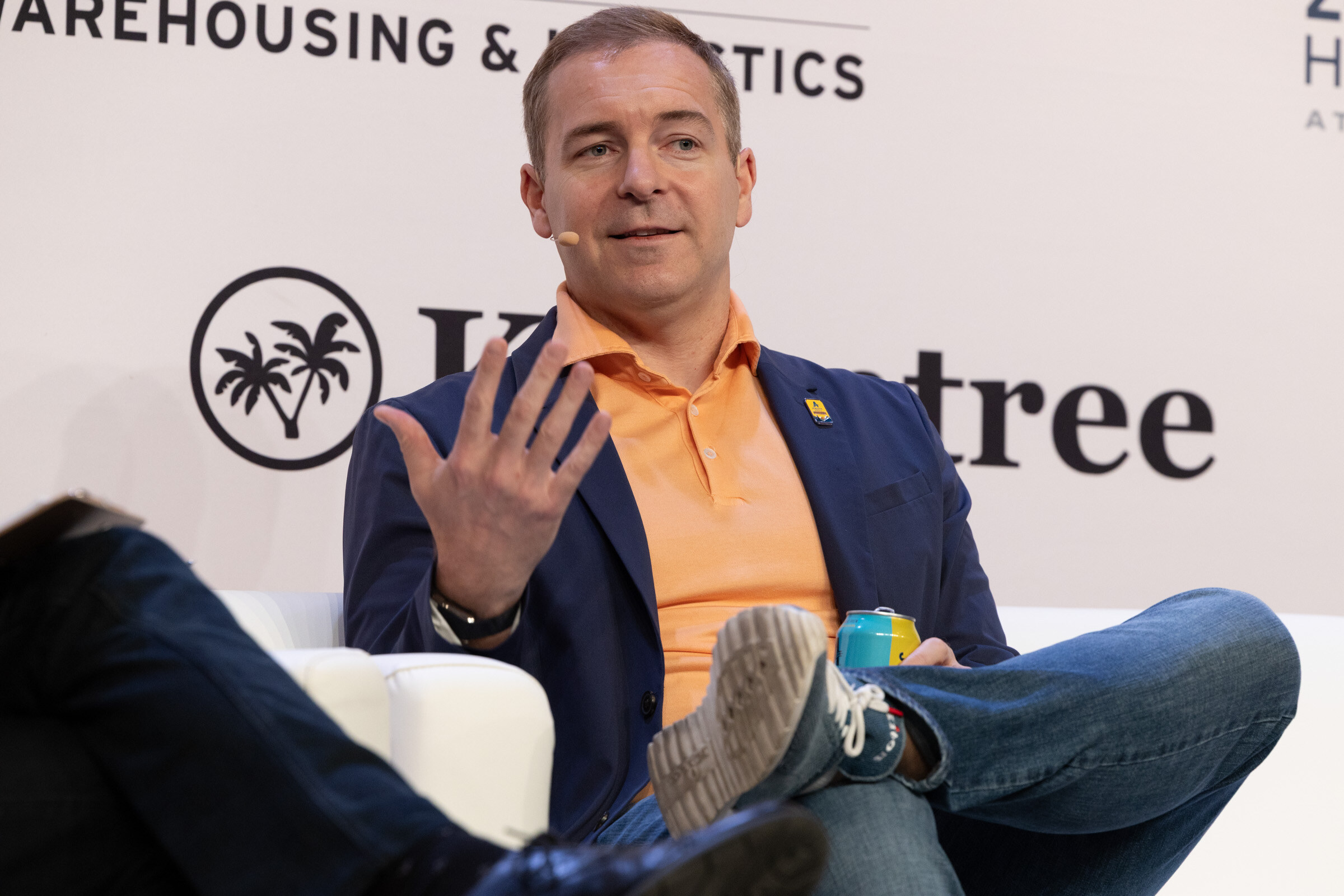 Man in an orange polo and navy blazer gesturing as he speaks, legs crossed, holding a can.