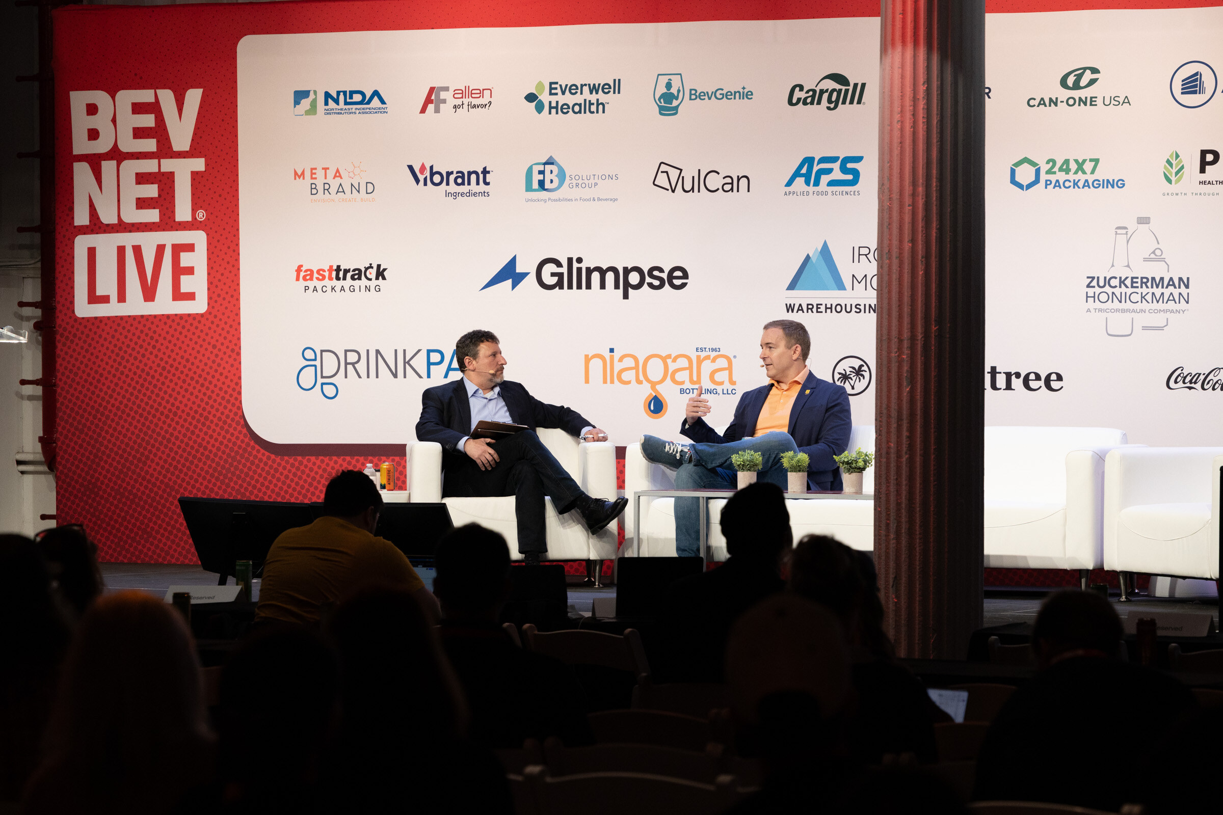 Two men seated on stage in front of a red BevNET Live banner and sponsor wall, with the audience in shadow.