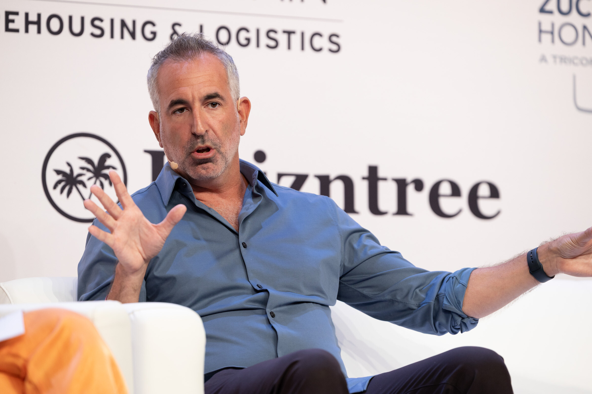 Man in a blue shirt gesturing with both hands as he speaks from a white couch in front of a Kaizntree logo.