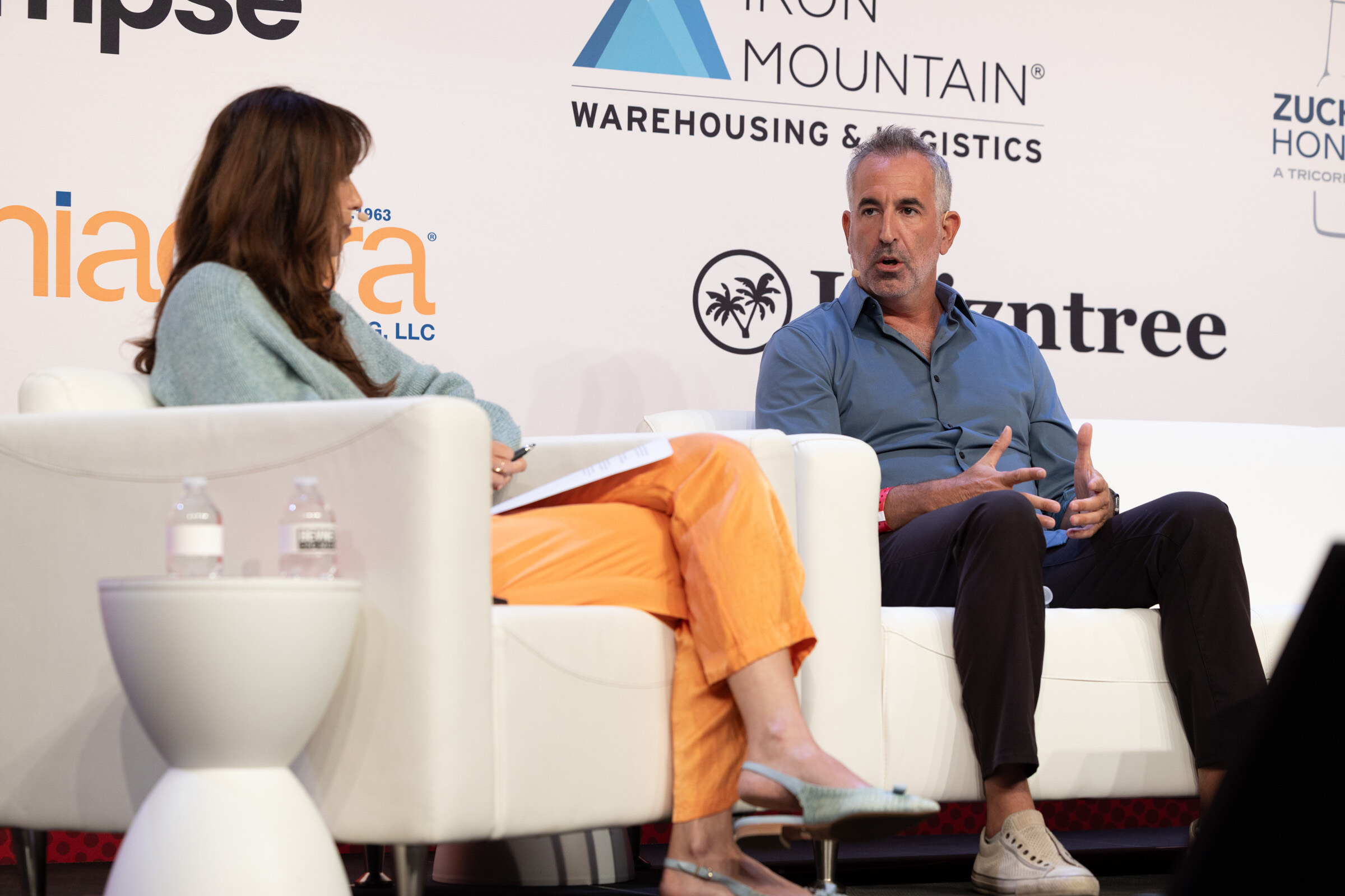 Woman in a sage sweater and orange trousers with notes facing a man in a blue shirt on white couches.