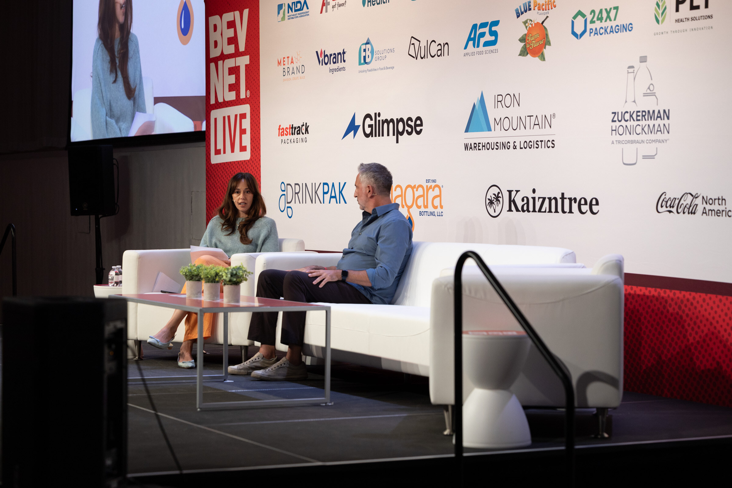 Two people seated on white couches on stage in front of a sponsor wall and a large screen.