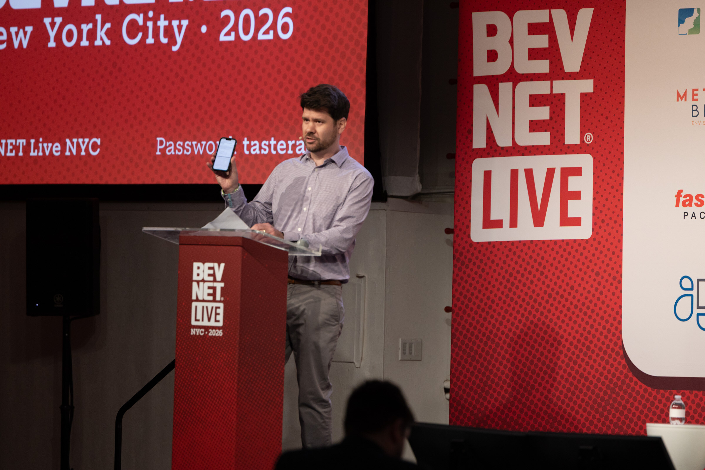 Man in a checked shirt at a red BevNET Live podium holding up a phone, in front of a red screen.