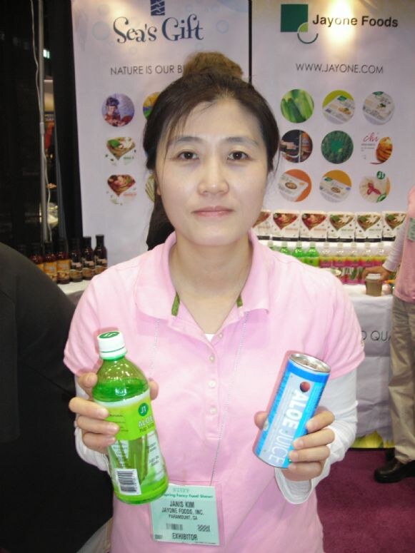 Janis Kim of Jayone Foods, Inc. in a pink polo holds a bottle of aloe drink and a can of aloe juice in front of Sea's Gift and Jayone Foods booth banners.
