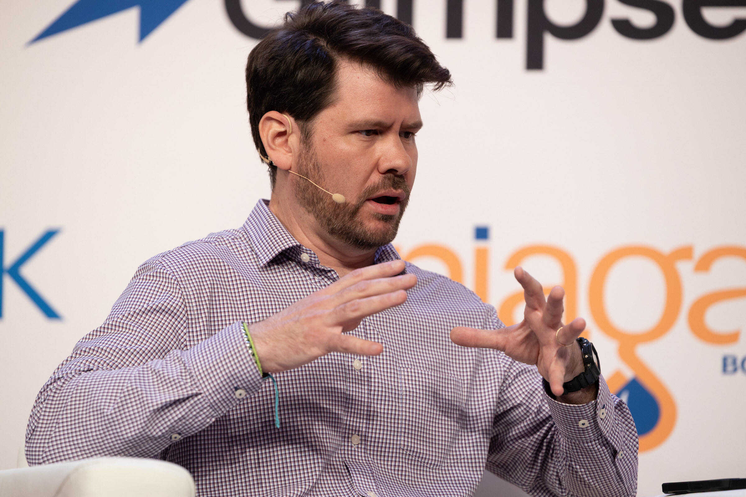 Man in a checked shirt with a headset microphone gesturing with both hands as he speaks.