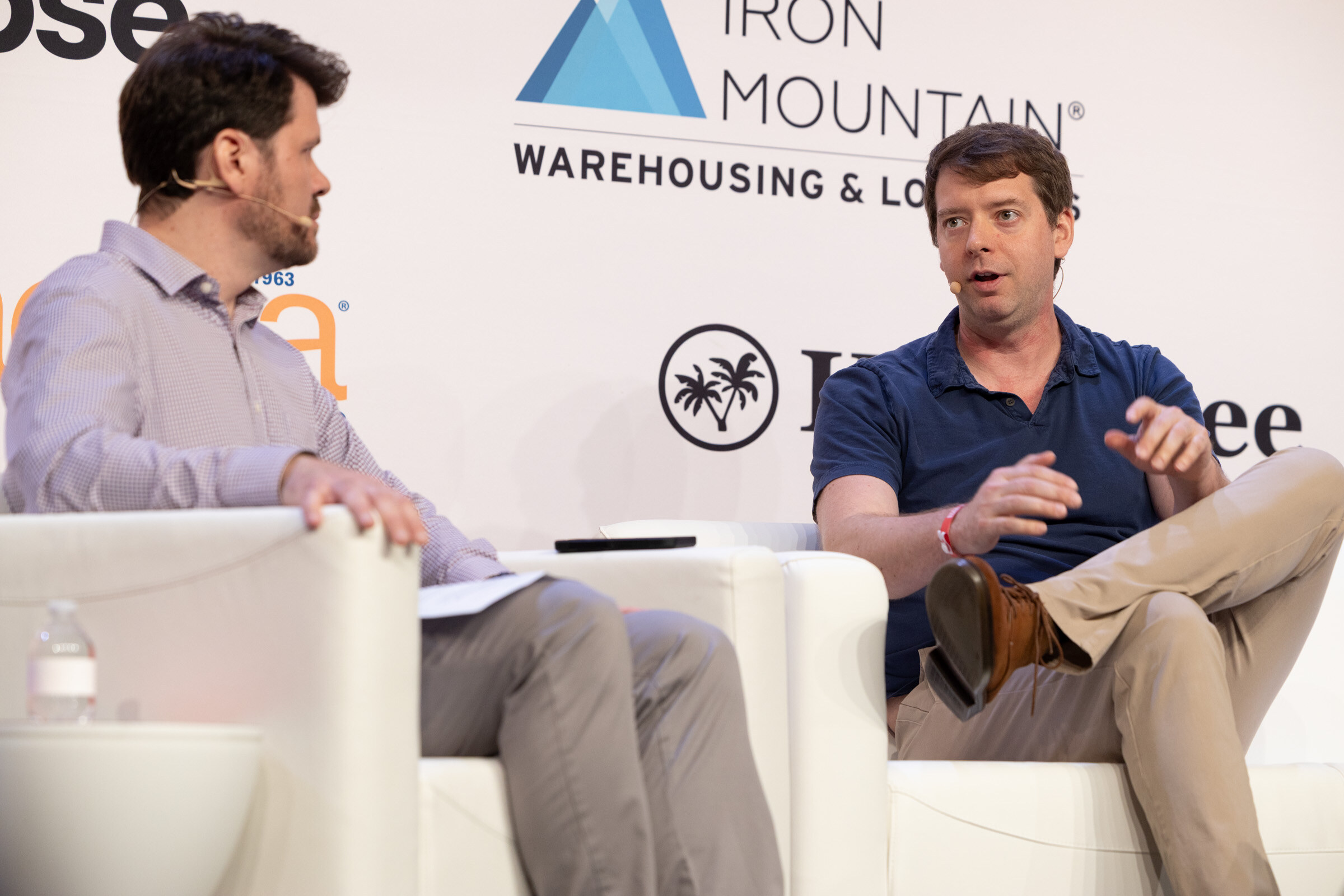 Man in a navy polo speaking and gesturing beside another man listening, in front of an Iron Mountain logo.